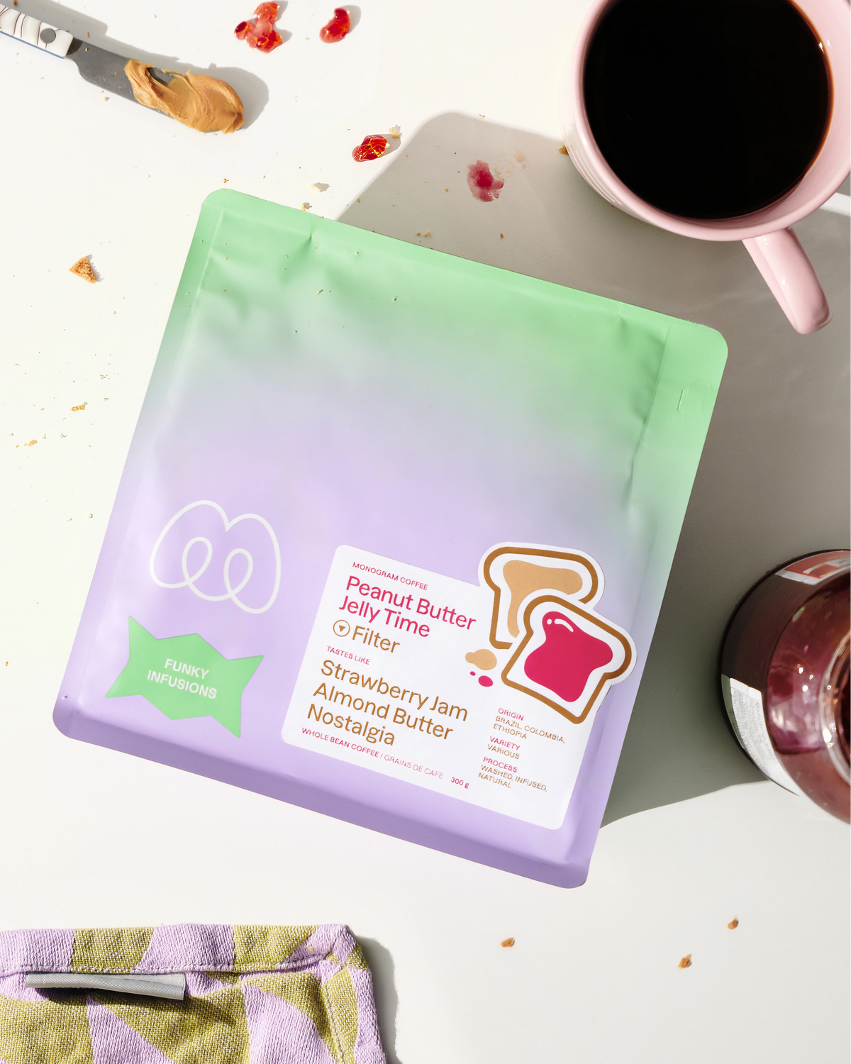 A purple and mint green gradient bag of Monogram Peanut Butter Jelly Time filter coffee on a sunny table with almond butter, toast graphics, and a pink mug, featuring tasting notes of strawberry jam, almond butter, and nostalgia.