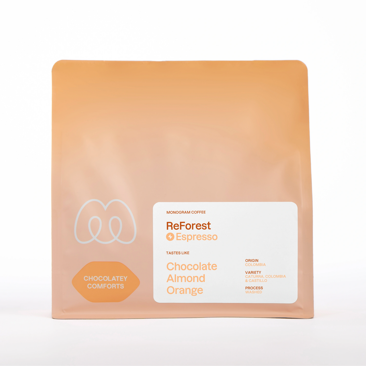 A warm beige and tan gradient bag of Monogram ReForest coffee, a washed processed Caturra, Castillo, and Colombia variety blend from Colombia with a Chocolatey Comforts label badge and tasting notes of chocolate, almond, and orange.