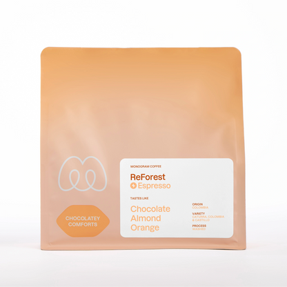 A warm beige and tan gradient bag of Monogram ReForest coffee, a washed processed Caturra, Castillo, and Colombia variety blend from Colombia with a Chocolatey Comforts label badge and tasting notes of chocolate, almond, and orange.