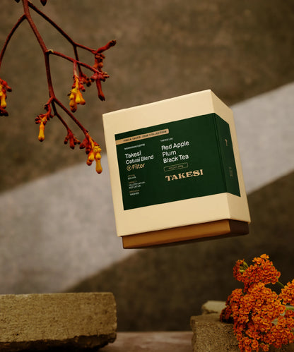 A cream and dark forest green box of Monogram Takesi Catuai Blend filter coffee floating in mid-air, a washed specialty coffee from Bolivia with tasting notes of red apple, plum, and black tea.
